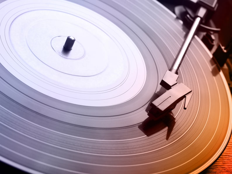 Op de afbeelding: Close-up van een vintage platenspeler met een vinylplaat. De plaat is grijs met concentrische groeven en de toonarm is boven de plaat geplaatst. De afbeelding heeft een warme, oranje en paarse gradi&euml;nt.