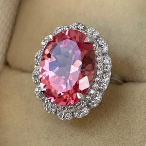 May include: A silver ring featuring a large, oval-cut pink gemstone surrounded by a halo of smaller, clear gemstones. The ring's setting is detailed, and the pink stone has multiple facets.