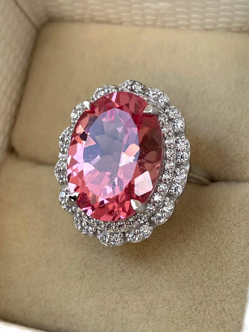 May include: A silver ring featuring a large, oval-cut pink gemstone surrounded by a halo of smaller, clear gemstones. The ring's setting is detailed, and the pink stone has multiple facets.