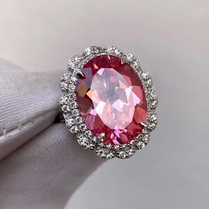 May include: A large, oval-cut pink gemstone ring with a halo of small, clear gemstones. The ring's setting is silver-colored, and the gemstones are set closely together. The pink gemstone has a faceted surface, reflecting light. The ring is displayed on a light gray surface.