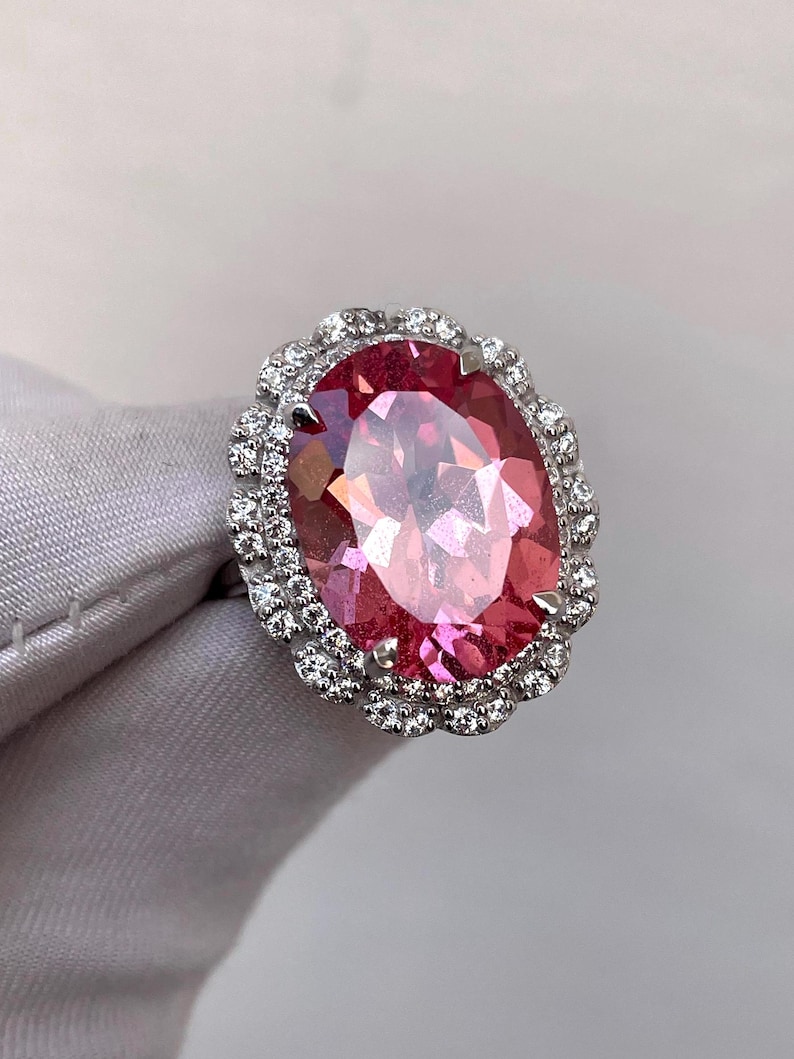 May include: A large, oval-cut pink gemstone ring with a halo of small, clear gemstones. The ring's setting is silver-colored, and the gemstones are set closely together. The pink gemstone has a faceted surface, reflecting light. The ring is displayed on a light gray surface.