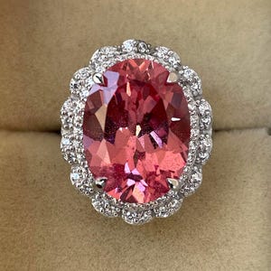 May include: A large, oval-cut pink gemstone ring with a halo of small, clear gemstones. The ring's setting is silver-colored, and the pink gemstone has multiple facets. The ring is set against a neutral background.