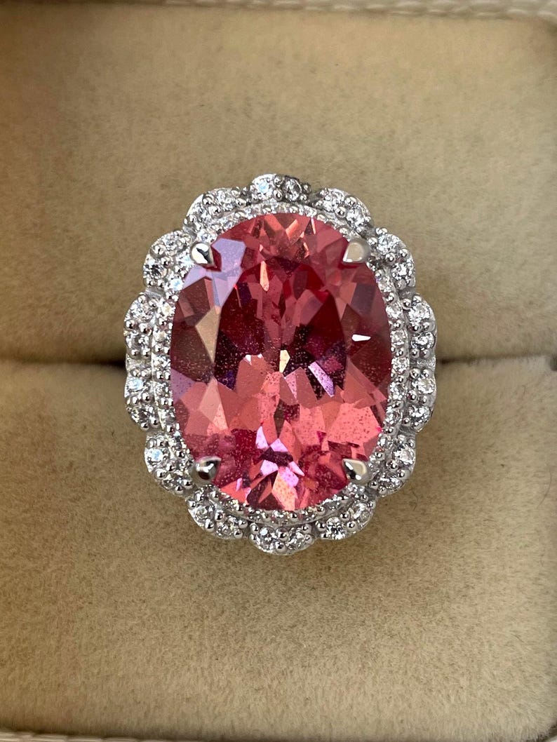 May include: A large, oval-cut pink gemstone ring with a halo of small, clear gemstones. The ring's setting is silver-colored, and the pink gemstone has multiple facets. The ring is set against a neutral background.