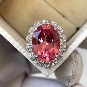 May include: A large, oval-cut pink gemstone ring with a halo of small, clear gemstones. The ring is set in a silver or white metal and is displayed in a white jewelry box. The gemstone has a vibrant, reddish-pink hue.
