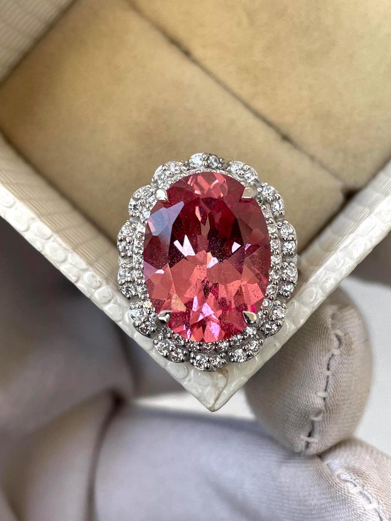 May include: A large, oval-cut pink gemstone ring with a halo of small, clear gemstones. The ring is set in a silver or white metal and is displayed in a white jewelry box. The gemstone has a vibrant, reddish-pink hue.