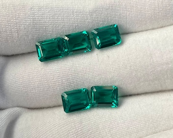 May include: Five emerald green rectangular gemstones, cut in a step-cut style, are arranged in two rows on a white fabric surface.
