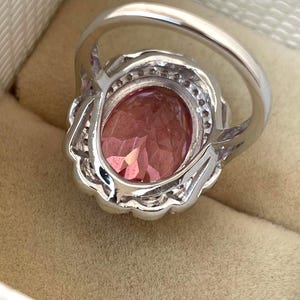May include: A silver ring with a large, oval-shaped pink gemstone. The gemstone is surrounded by smaller, clear stones and set in a decorative silver band. The ring is displayed in a beige jewelry box.