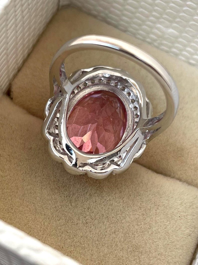 May include: A silver ring with a large, oval-shaped pink gemstone. The gemstone is surrounded by smaller, clear stones and set in a decorative silver band. The ring is displayed in a beige jewelry box.