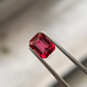 Può includere: Una pietra preziosa di colore rosso intenso con taglio rettangolare a smeraldo, tenuta da una pinzetta.