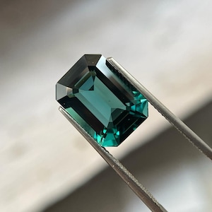 Peut inclure: Une pierre précieuse de couleur vert foncé taillée en émeraude, tenue par des pinces. La pierre précieuse est facetée et a une couleur profonde et riche.