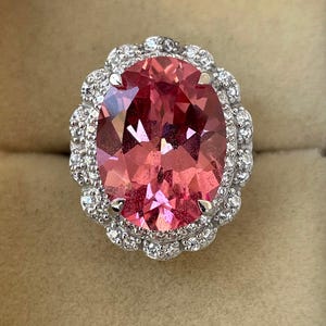 May include: An oval-shaped pink gemstone ring with a halo of small, clear gemstones. The ring's setting is silver-colored, and the gemstones are set closely together, creating a sparkling effect. The ring is displayed against a neutral background.