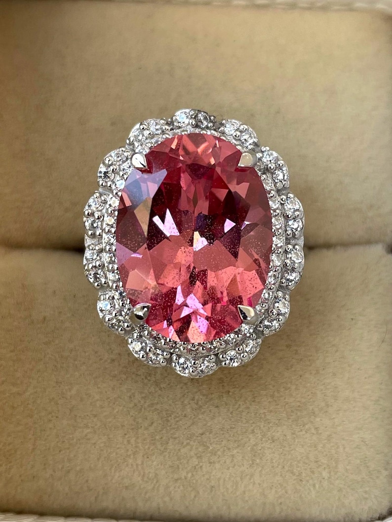 May include: An oval-shaped pink gemstone ring with a halo of small, clear gemstones. The ring's setting is silver-colored, and the gemstones are set closely together, creating a sparkling effect. The ring is displayed against a neutral background.