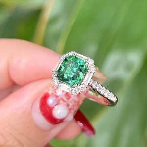 Pu&ograve; includere: Un anello in oro bianco con una pietra preziosa verde di forma quadrata circondata da un alone di diamanti pi&ugrave; piccoli. L'anello &egrave; incastonato in oro bianco e ha una fascia delicata.