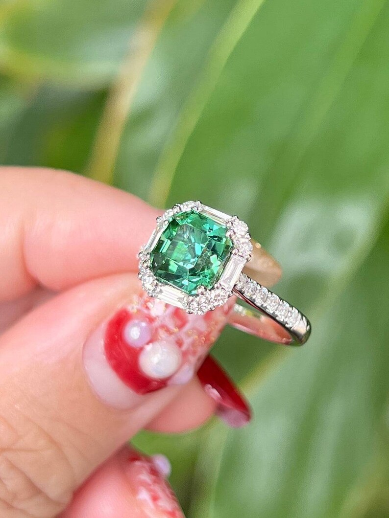 Pu&ograve; includere: Un anello in oro bianco con una pietra preziosa verde di forma quadrata circondata da un alone di diamanti pi&ugrave; piccoli. L'anello &egrave; incastonato in oro bianco e ha una fascia delicata.