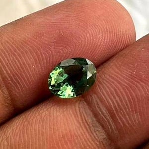 Op de afbeelding: Een gefacetteerde, ovaalvormige edelsteen met een diepgroene tint. De edelsteen vertoont een briljante slijpvorm, die het licht reflecteert en de interne helderheid laat zien. De steen wordt tussen vingers gehouden, wat de grootte en kleur benadrukt.
