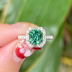 Pu&ograve; includere: Un anello con una pietra preziosa verde di forma quadrata con un alone di diamanti pi&ugrave; piccoli. L'anello &egrave; incastonato in argento.