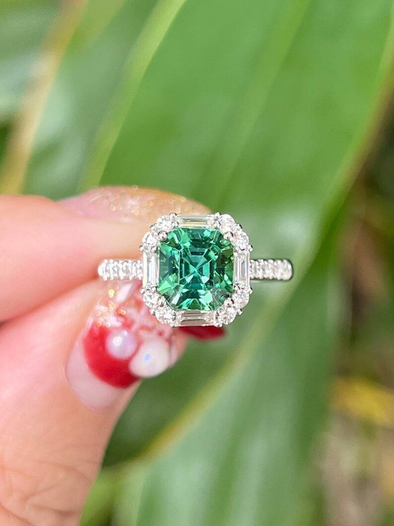 Pu&ograve; includere: Un anello con una pietra preziosa verde di forma quadrata con un alone di diamanti pi&ugrave; piccoli. L'anello &egrave; incastonato in argento.