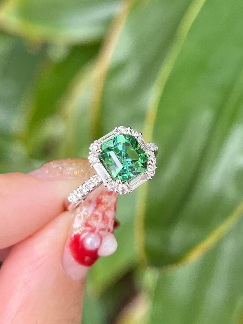 Pu&ograve; includere: Un anello d'argento con una pietra preziosa verde circondata da un'aureola di diamanti pi&ugrave; piccoli.