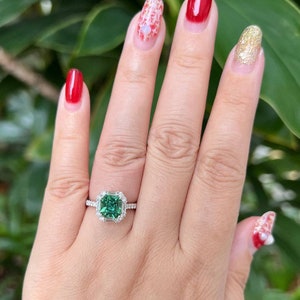 Pu&ograve; includere: Primo piano di una mano che indossa un anello con una grande pietra preziosa verde di forma quadrata circondata da diamanti pi&ugrave; piccoli. L'anello &egrave; in metallo bianco. La mano indossa anche lo smalto per unghie di vari colori, tra cui rosso, bianco e oro.