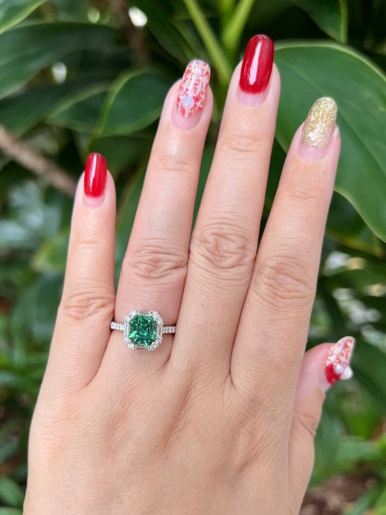 Pu&ograve; includere: Primo piano di una mano che indossa un anello con una grande pietra preziosa verde di forma quadrata circondata da diamanti pi&ugrave; piccoli. L'anello &egrave; in metallo bianco. La mano indossa anche lo smalto per unghie di vari colori, tra cui rosso, bianco e oro.