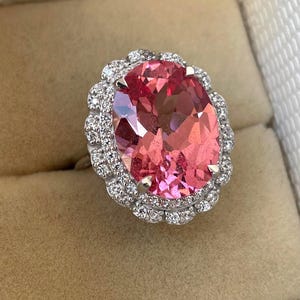 May include: A large, oval-cut pink gemstone ring with a halo of small, clear gemstones. The ring's band is silver-colored, and the setting is intricate. The ring is displayed in a beige jewelry box.
