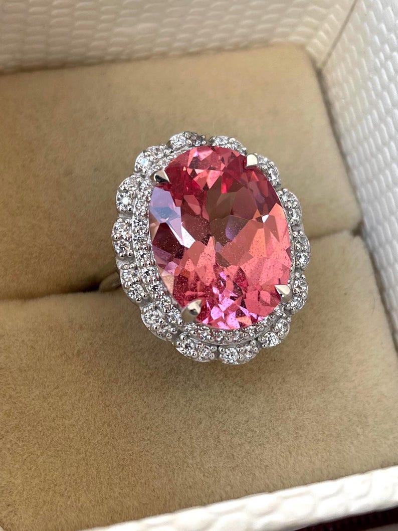 May include: A large, oval-cut pink gemstone ring with a halo of small, clear gemstones. The ring's band is silver-colored, and the setting is intricate. The ring is displayed in a beige jewelry box.