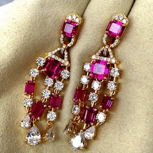 以下が含まれることがあります： ピンク色の長方形と洋梨型の宝石と、透明な丸い石が交互に配置された、金色のイヤリングのペア。 イヤリングは、詳細で装飾的なデザインです。