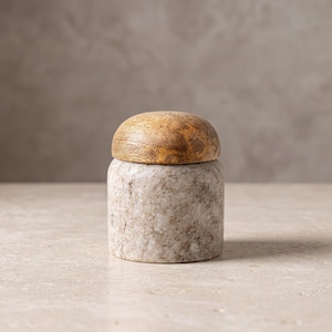 Puede incluir: Un recipiente cilíndrico con una base de piedra moteada en tonos beige y gris, rematado por una tapa de madera natural lisa y redondeada. El cuerpo de piedra muestra una textura granulada, mientras que la tapa presenta vetas visibles.