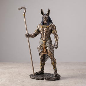Puede incluir: Una estatua de tono bronce de la deidad egipcia Anubis sobre una base con textura de piedra. La figura tiene cabeza de chacal, torso humano musculoso y viste un taparrabos drapeado con detalles dorados. Sostiene un bastón rematado con una cobra.