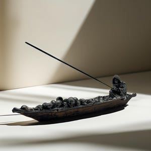 Könnte beinhalten: Schwarzer Räucherofen in Form eines Bootes, gefüllt mit Schädeln, mit einer Kapuzenfigur, die am Bug sitzt. Der Räucherstäbchen wird in die Hand der Figur gelegt.