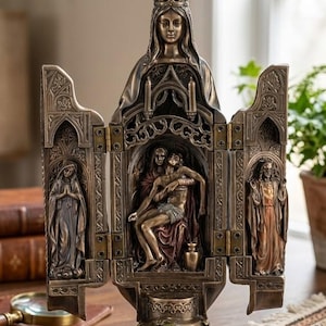 May include: A bronze-toned religious triptych sculpture featuring a central scene of a figure being held by another, flanked by figures in relief. The sculpture has a crown at the top and is set on a decorative base with a serpent.