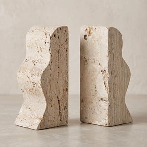 Puede incluir: Un par de sujetalibros de travertino beige con una textura porosa natural y una silueta ondulada y curva distintiva en un lado, con un borde vertical plano en el otro, mostrando las variaciones orgánicas y los tonos tierra de la piedra.