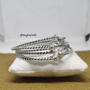 Puede incluir: Una pulsera plateada con un diseño de cuerda trenzada. La pulsera está compuesta por múltiples bandas, cada una con una apariencia texturizada y trenzada. Pequeñas cuentas decorativas plateadas se colocan a intervalos a lo largo de la pulsera. La pulsera descansa sobre un cojín blanco.