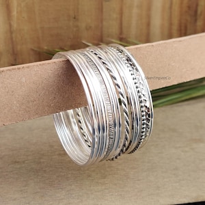 Puede incluir: Un conjunto de nueve pulseras de plata con diferentes texturas y patrones. Las pulseras están apiladas una encima de la otra y están dispuestas en forma circular. Las pulseras están hechas de plata de ley y tienen un acabado pulido.