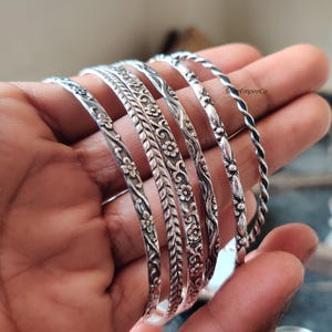 Könnte beinhalten: Eine Sammlung silberner Armreifen mit aufwendigen Blumen- und Blattmustern. Die Armreifen variieren im Design, einige weisen gedrehte Seiltexturen auf. Der Schmuck wird auf einer Hand präsentiert, um die Details jedes Stücks hervorzuheben.