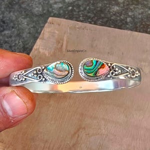 Puede incluir: Una pulsera de plata con dos incrustaciones ovaladas de concha de abulón. Las conchas iridiscentes están enmarcadas por un borde de cuentas y filigranas de plata decorativas. La pulsera está abierta en la parte inferior.