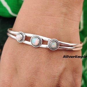 May include: Silver bangle bracelet with three white opal stones set in silver bezels. The bracelet is double-banded and has a delicate design.