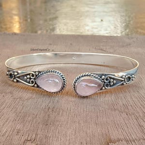 Puede incluir: Un brazalete de plata con dos piedras de cuarzo rosa. El brazalete tiene un diseño delicado con pequeños detalles florales.