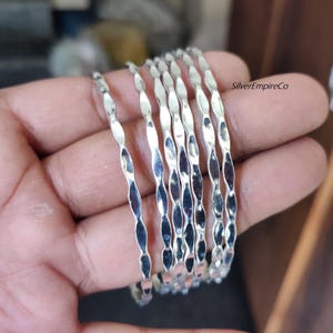 Könnte beinhalten: Ein Satz von sieben silberfarbenen Armreifen. Jeder Armreif hat ein verdrehtes, längliches ovales Design. Die Armreifen sind gestapelt und reflektieren das Licht. Der Schmuck wird vor einem neutralen Hintergrund präsentiert.