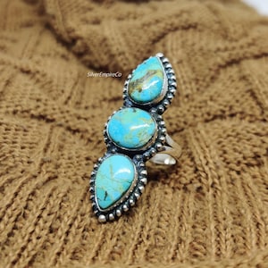 May include: A silver ring with three turquoise stones set in a vertical row. The stones are a teardrop shape and a round shape. The ring is on a brown knitted fabric.