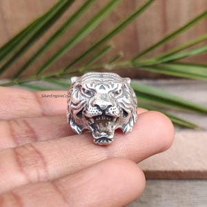 May include: A silver ring with a detailed tiger head design. The tiger's face is sculpted with open jaws, showing teeth and intricate fur patterns. The ring is held by a person's fingers, with the brand name "SilverEmpireCo" visible.