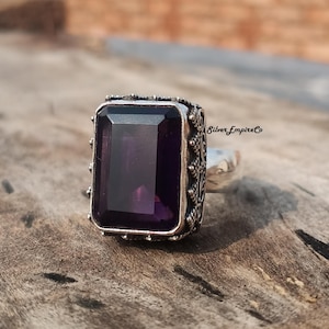 以下が含まれることがあります： 大きな長方形の濃い紫色の宝石をあしらったシルバーリング。リングには宝石の周りとバンドに装飾的なディテールがあります。「Silver Empire Co」の文字が側面に表示されています。