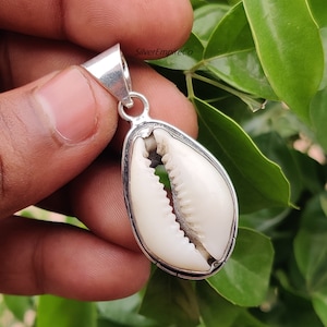 May include: A silver pendant featuring a white cowrie shell. The pendant has a bail for attaching to a chain or cord.