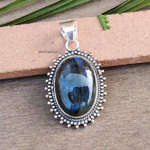 May include: A silver pendant with a large oval-shaped labradorite gemstone set in a detailed silver frame. The gemstone has a dark blue and green iridescent sheen.