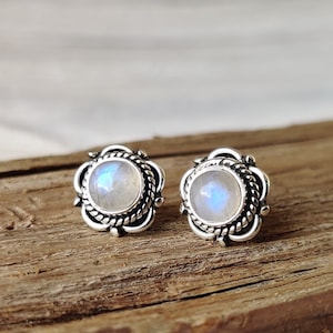 May include: A pair of silver earrings featuring a round moonstone cabochon set in a decorative silver bezel. The earrings have a floral design.