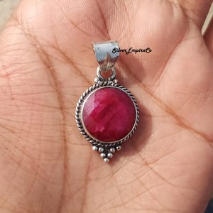 May include: A round, ruby-colored pendant with a silver-colored bail. The pendant has a textured silver border and a cluster of small silver beads at the bottom. The text "Silver Empire Co" is visible on the bail.
