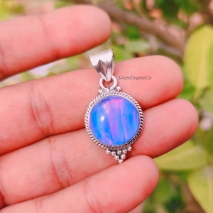 Op de afbeelding: Een zilveren hanger met een ronde, blauwe en roze opaal edelsteen. De hanger heeft een decoratieve rand in touwstijl en een kleine lus voor een ketting. De achtergrond is wazig, met groene bladeren en een hand die de hanger vasthoudt.