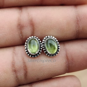 Puede incluir: Un par de pendientes de plata con piedras preciosas ovaladas verdes engastadas en un borde de cuentas. Los pendientes se muestran en la mano de una persona.