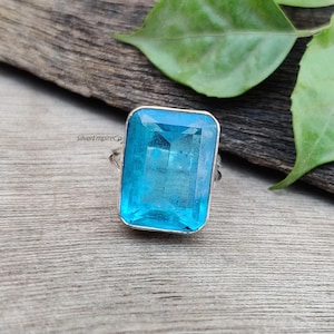 Puede incluir: Un anillo de plata con una gran piedra rectangular de color azul claro. El anillo está colocado sobre una superficie de madera.