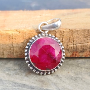 May include: A round, silver pendant with a large, faceted, ruby-red gemstone. The gemstone is surrounded by a beaded silver border. The pendant has a silver loop at the top for a chain. The pendant is displayed on a wooden surface.